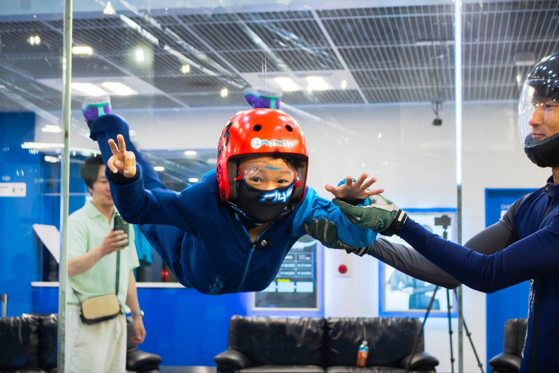赤いヘルメットと青いスーツを着た子供が、インストラクターに支えられながらインドアスカイダイビングを楽しんでいる様子
