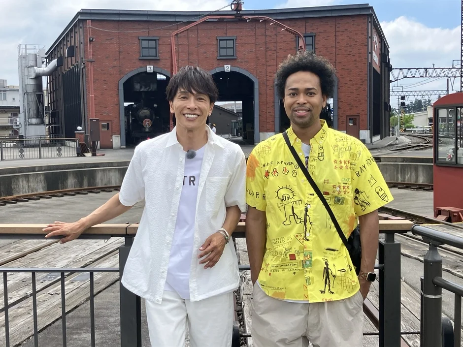 鉄道施設を背景に笑顔で並んで立つ2人の男性