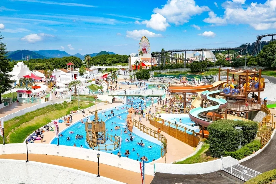晴れた夏の日に多くの人々で賑わう広々としたウォーターパークの全景です。流れるプールやウォータースライダー、海賊船の遊具があり、遠景には観覧車やジェットコースターのある遊園地も見えます。