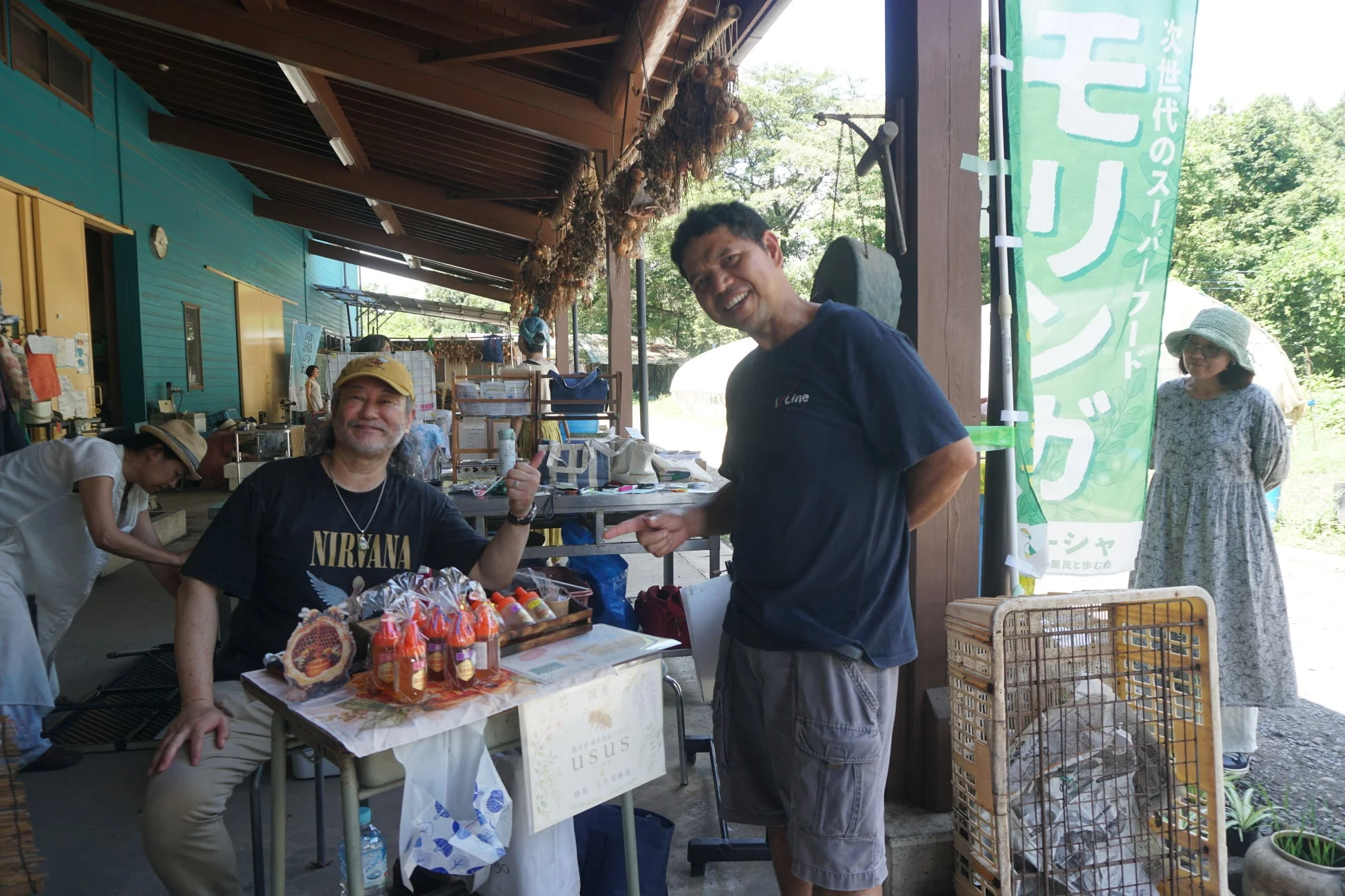 屋外の直売所で笑顔の男性二人と商品、モリンガののぼり