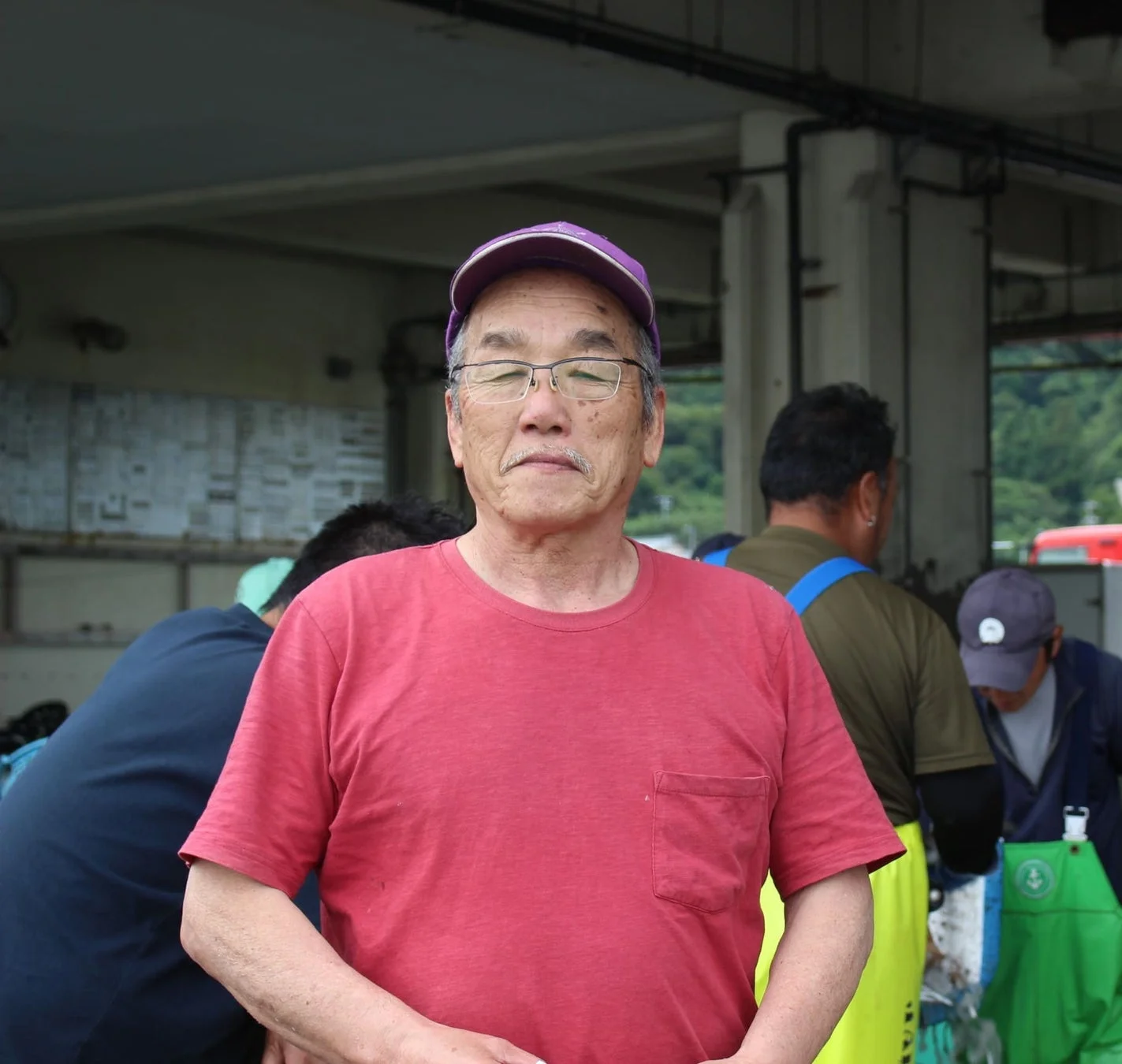 赤いTシャツを着て紫色の帽子と眼鏡をかけた高齢の男性がカメラを見つめています。背景には他の人々が作業している様子が見られ、市場のような活気ある場所が伺えます。