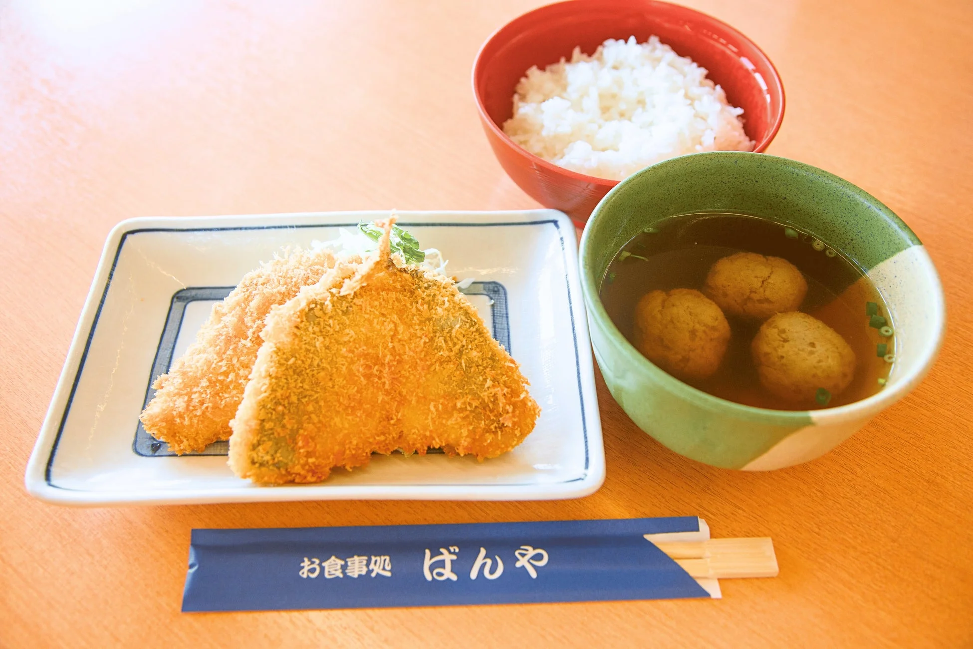 木製のテーブルに置かれた和食の定食セットです。サクサクに揚がったフライが2つ、白いご飯、そして団子が入った汁物が並べられています。箸袋には「お食事処 ばんや」と書かれており、日本の食堂での一食を捉えた写真です。