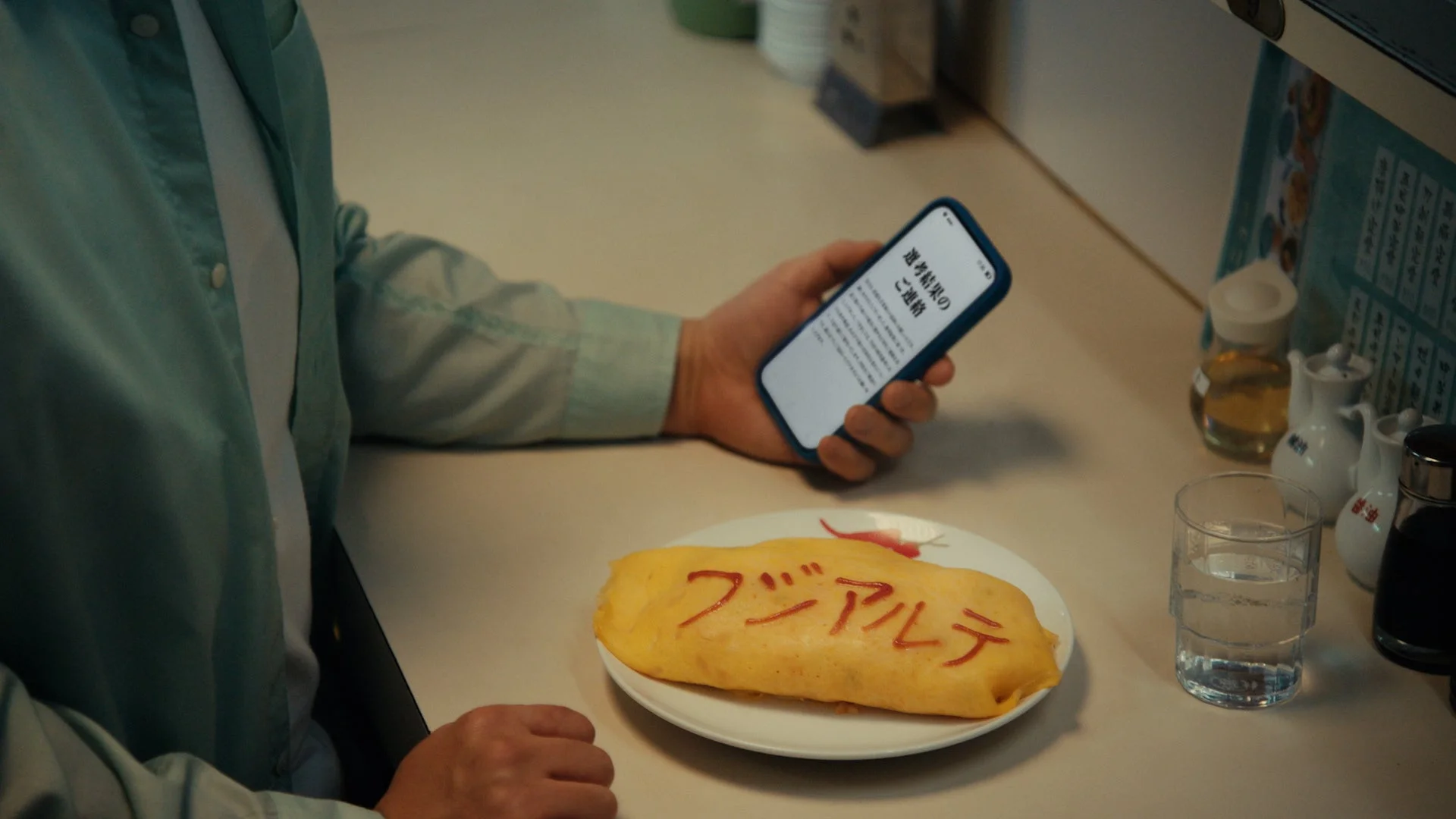 オムライスを前にスマートフォンを操作する人物のク様子。オムライスにはケチャップで「フシアルテ」と書かれ、スマホ画面には「選考結果のご連絡」と表示されており、食事中に重要な通知を確認している場面です。