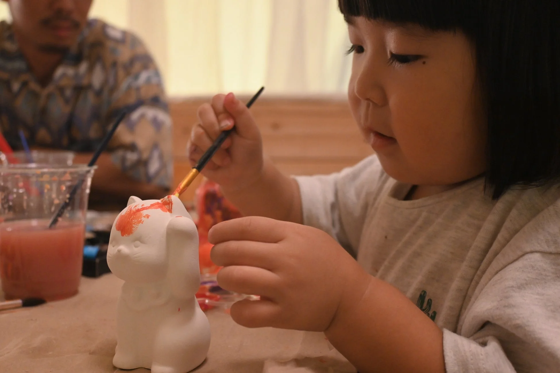 小さな子供が白い猫の陶器の置物にオレンジ色の絵の具で色を塗っている様子