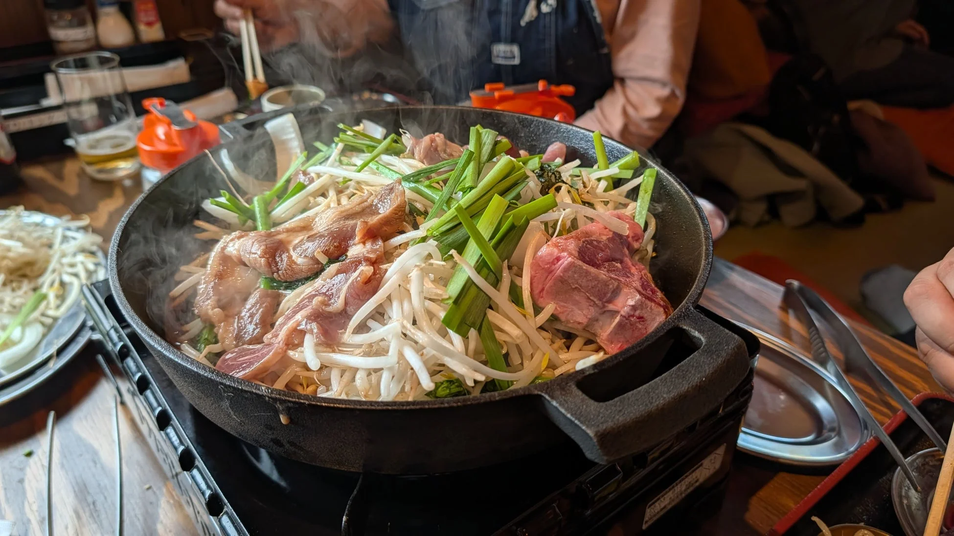 湯気が立ち上る鉄板で、羊肉とたっぷり野菜(もやし、ネギなど)を調理しているジンギスカンの食事風景です。