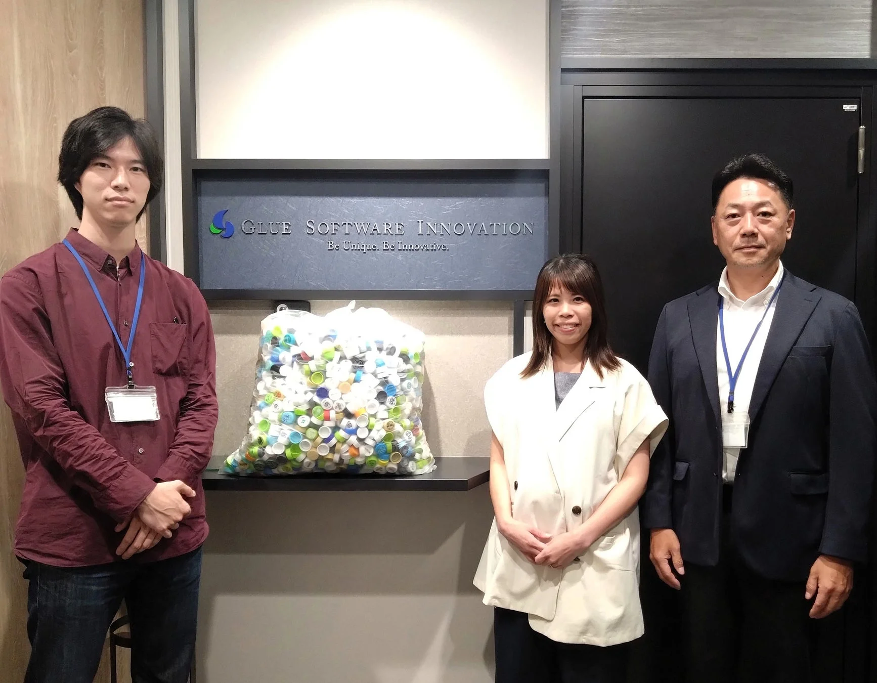 会社のオフィスで、3人の社員が「GLUE SOFTWARE INNOVATION」のロゴの前で立っています。棚には大量のカラフルなペットボトルキャップが入った袋が置かれており、環境活動やリサイクルへの取り組みを示唆しています。
