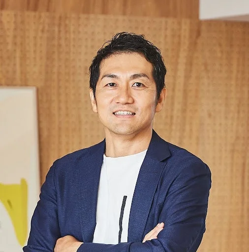 青いジャケットと白いTシャツを着たアジア人男性のポートレート。短髪で、カメラに向かって穏やかな笑顔を見せており、自信と親しみやすさを感じさせる。背景は木目調の壁です。