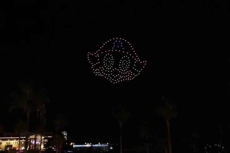 夜空にドローンで描かれた可愛らしいキャラクターの顔が浮かび上がっています。下にはヤシの木とイルミネーションで飾られた建物が見え、リゾート地での特別なイベントの様子を捉えた一枚です。