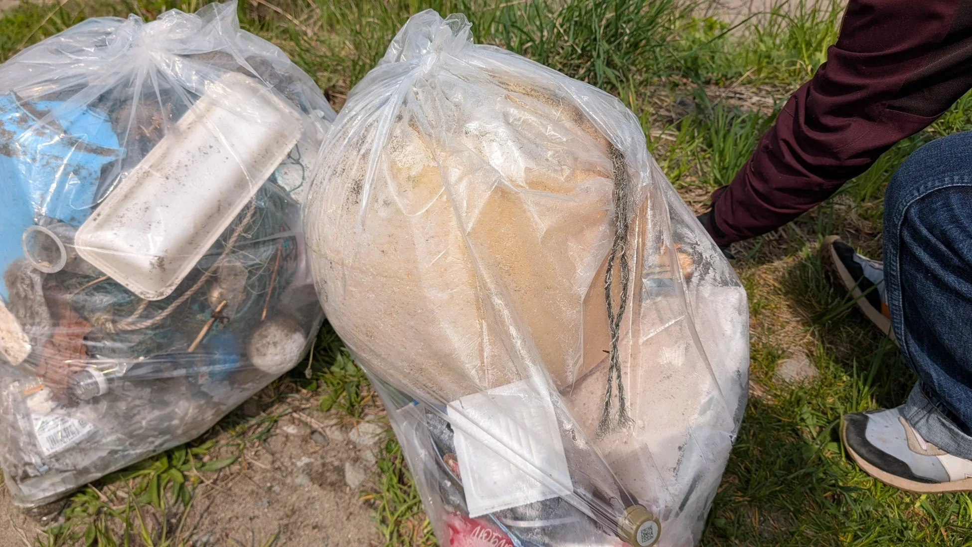 草地に置かれた透明なゴミ袋2つに、プラスチック片や砂など様々なゴミが詰められている。