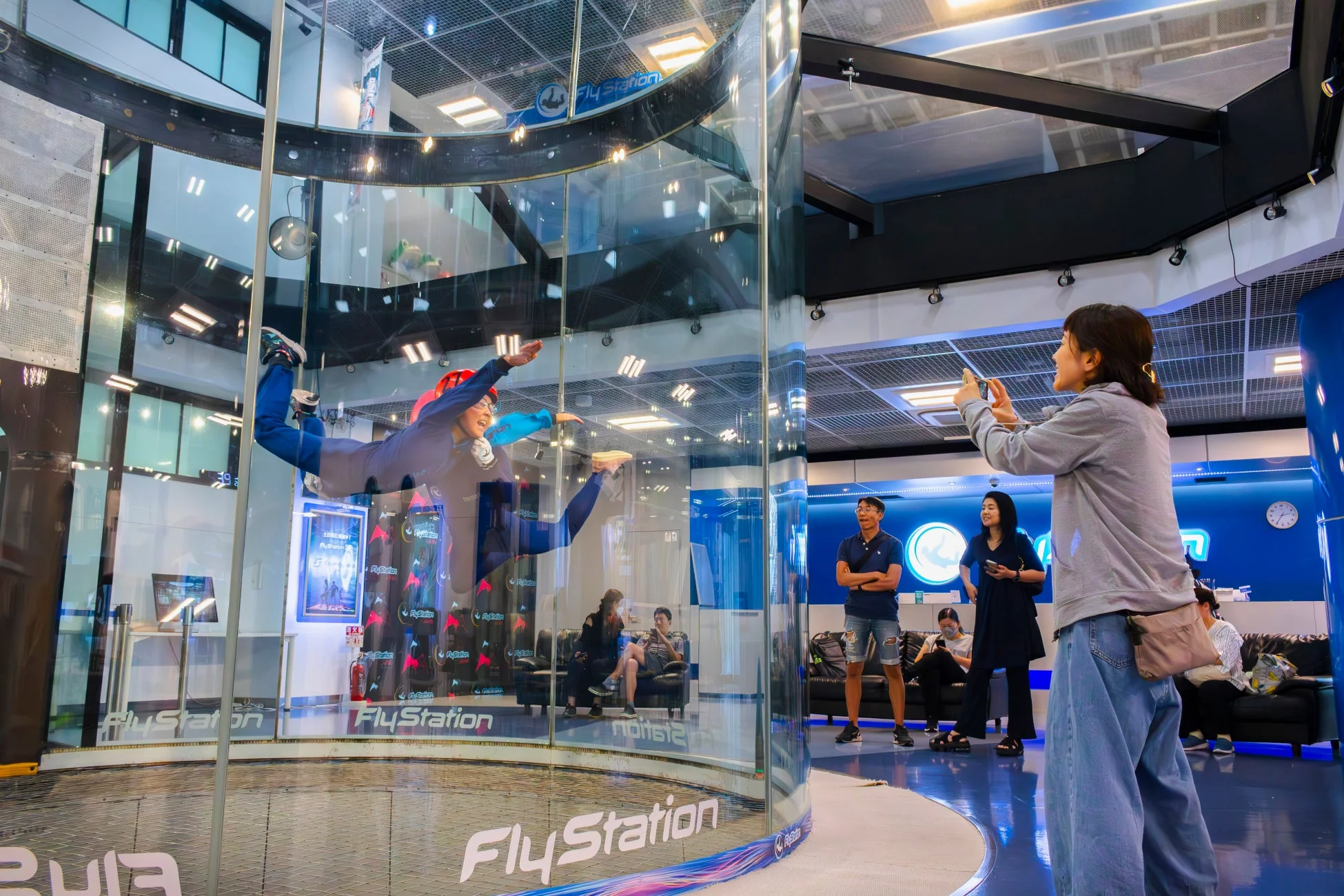 屋内スカイダイビング施設「FlyStation」で、フライトスーツを着た人がガラスのトンネル内で浮遊体験をしている様子