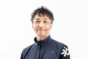 白背景の前で、ダークブルーのスポーツウェアを着た中年の男性が穏やかに微笑んでいるポートレート写真です。