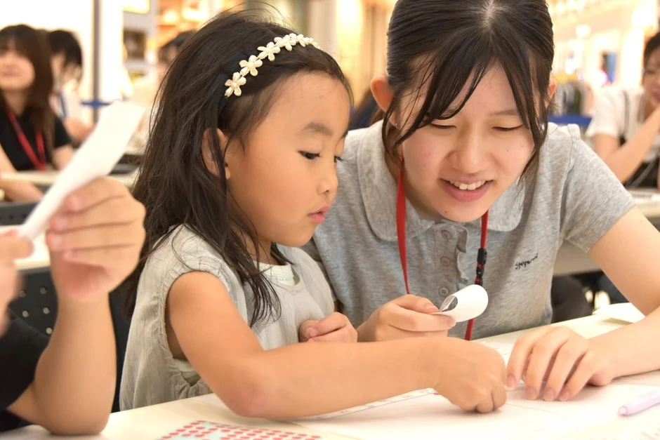 若い女性が小さな女の子に、紙を使った作業を教えている様子