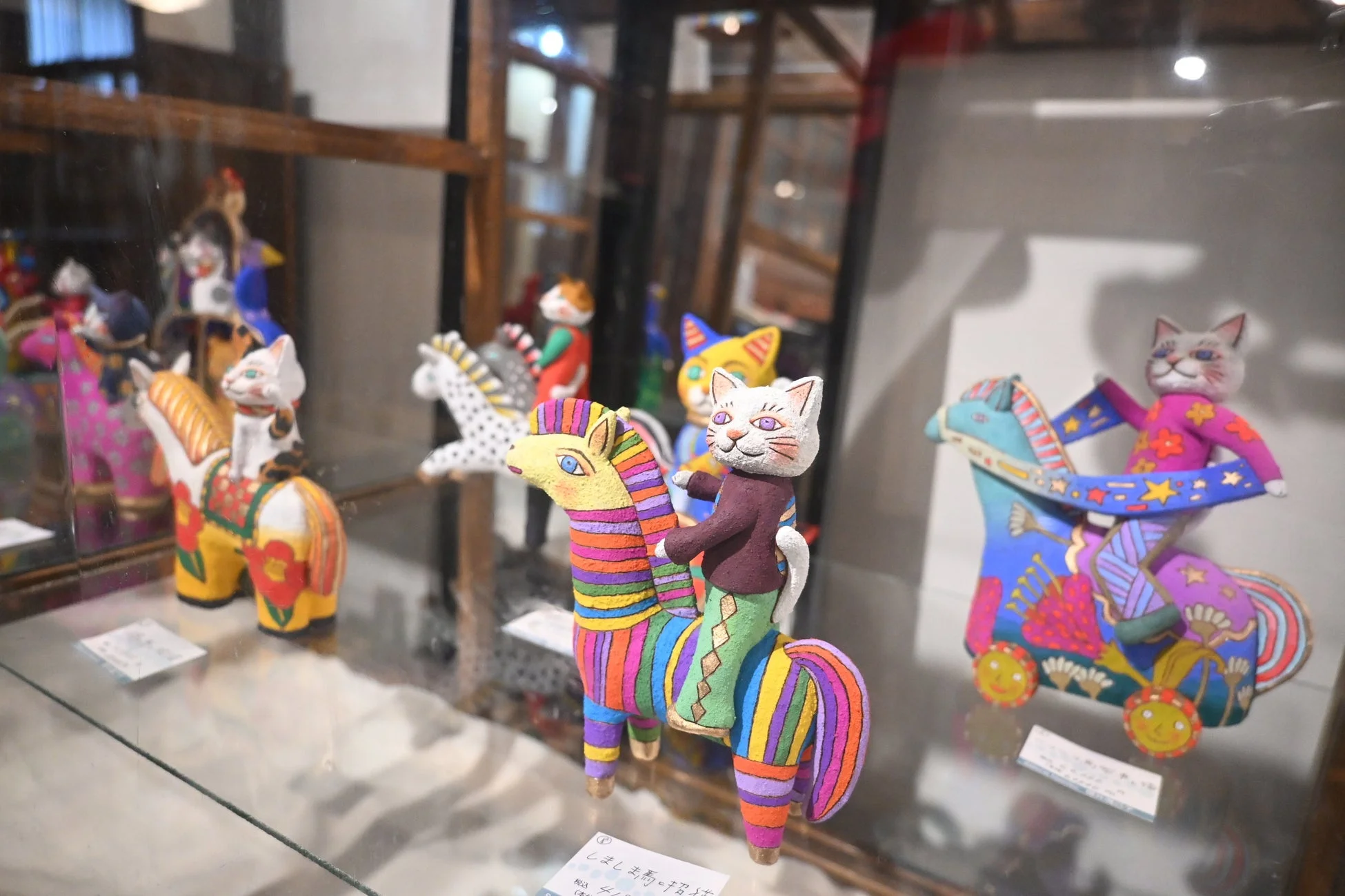 ガラスケースの中に、猫が馬に乗ったカラフルで個性的な置物が複数展示されています