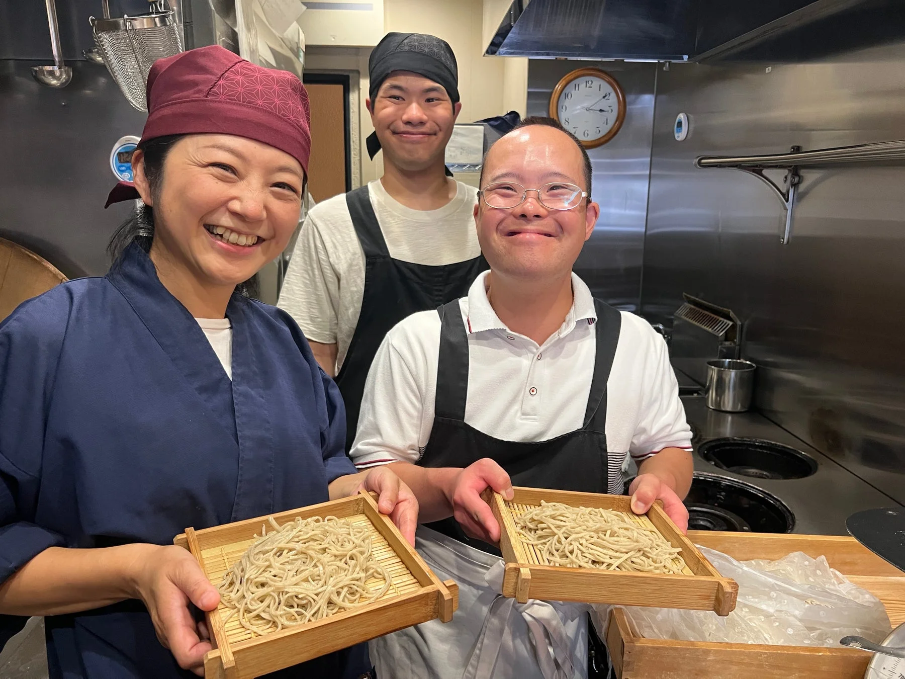 厨房で3人の人々が笑顔で並び、手作りのそばが入った木製のざるを2人が持っています。