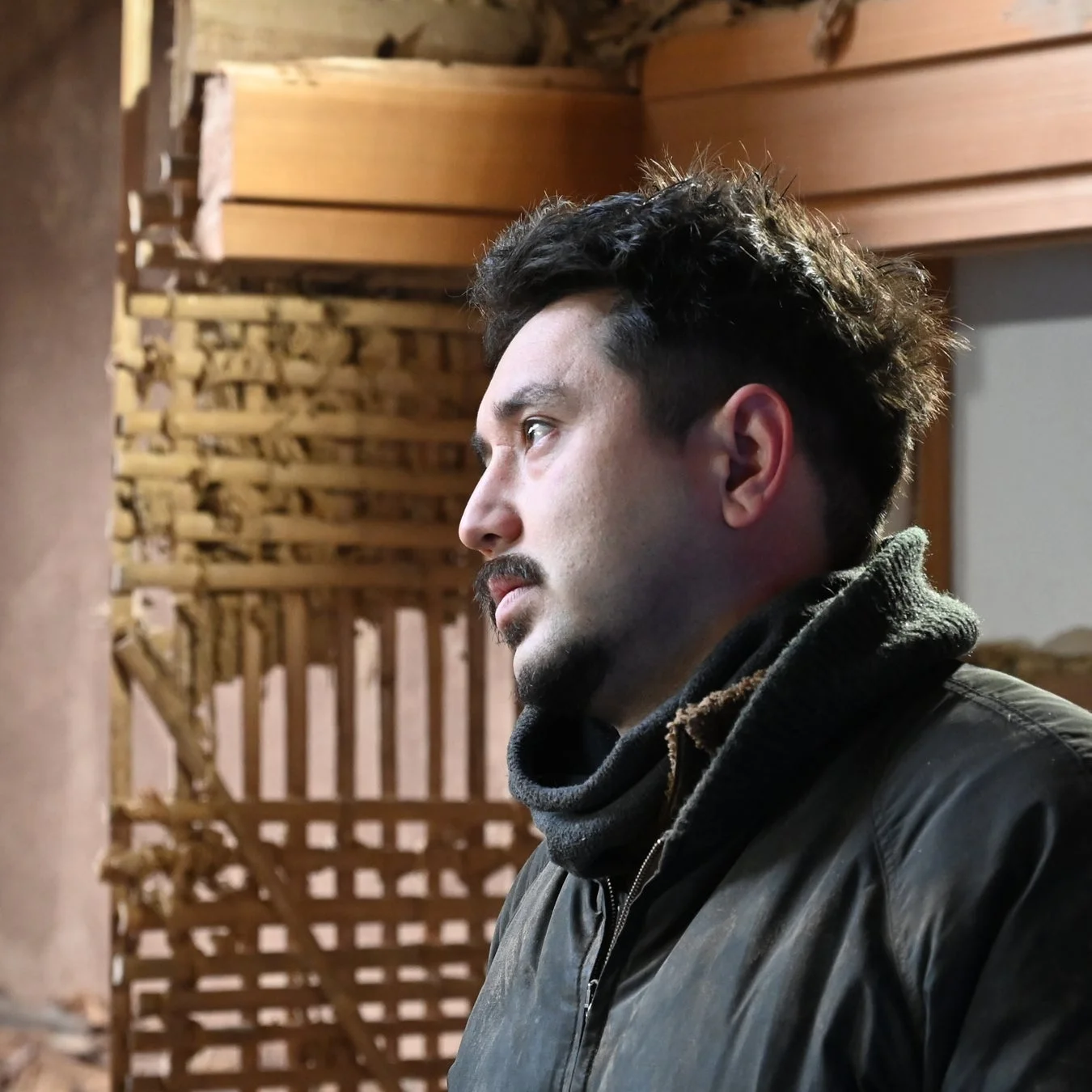 工事中または改装中の建物内で、横顔を見せる男性のポートレート。彼は黒いジャケットを着用し、真剣な表情で遠くを見つめています。背景にはむき出しの木材の構造が見えます。