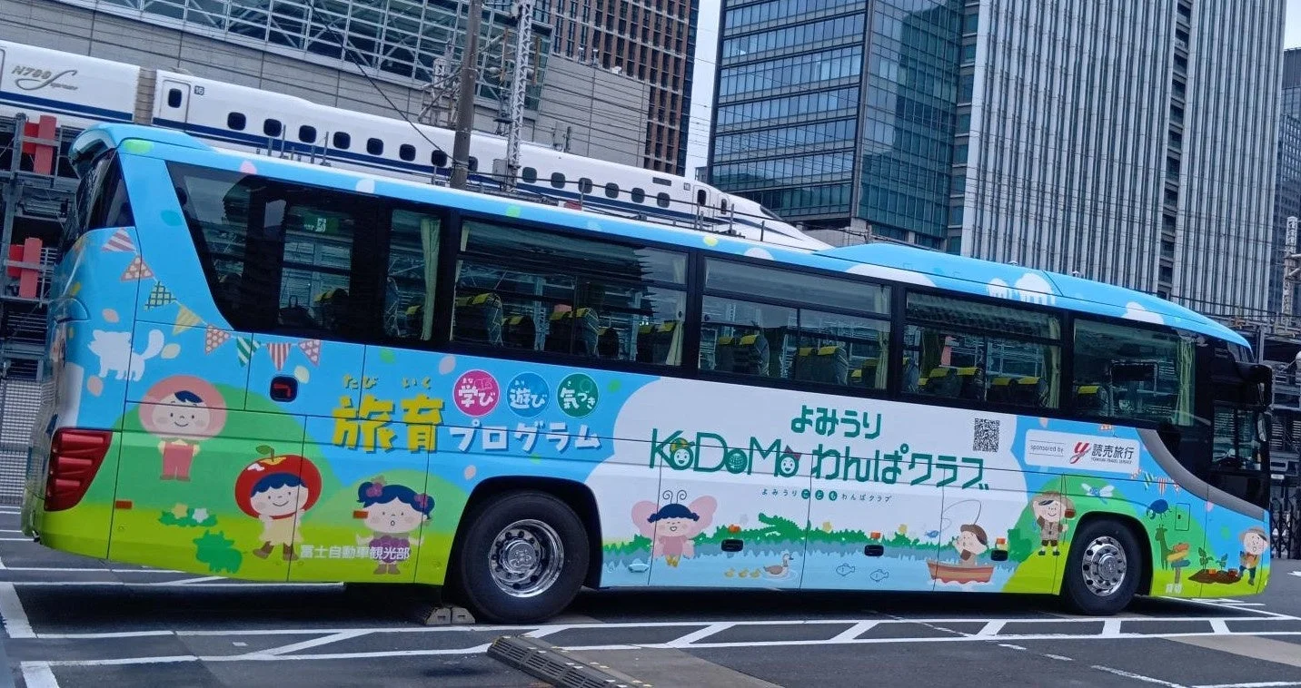 青と緑を基調とした、子供向けのキャラクターが描かれた「KoDoMo わんぱくクラス」の観光バスが、都市の背景に停車しています。上部には「N700」のロゴが見える新幹線が走っており、交通機関が並ぶ賑やかな風景です。