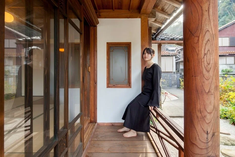 黒いドレスを着た若い女性が、日本の伝統的な木造家屋の縁側に座っているポートレート。静かで落ち着いた雰囲気の中で、和風建築の美しさが際立っています。