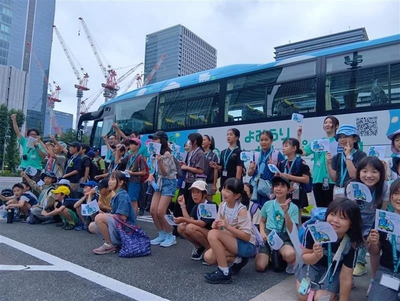 青いバスの前で、子供たちが楽しそうに集まっている様子。都市部の高層ビルや建設クレーンを背景に、多くの子供たちが笑顔で手を振ったり、うちわを持ったりしている。遠足かイベントの一コマと思われる。