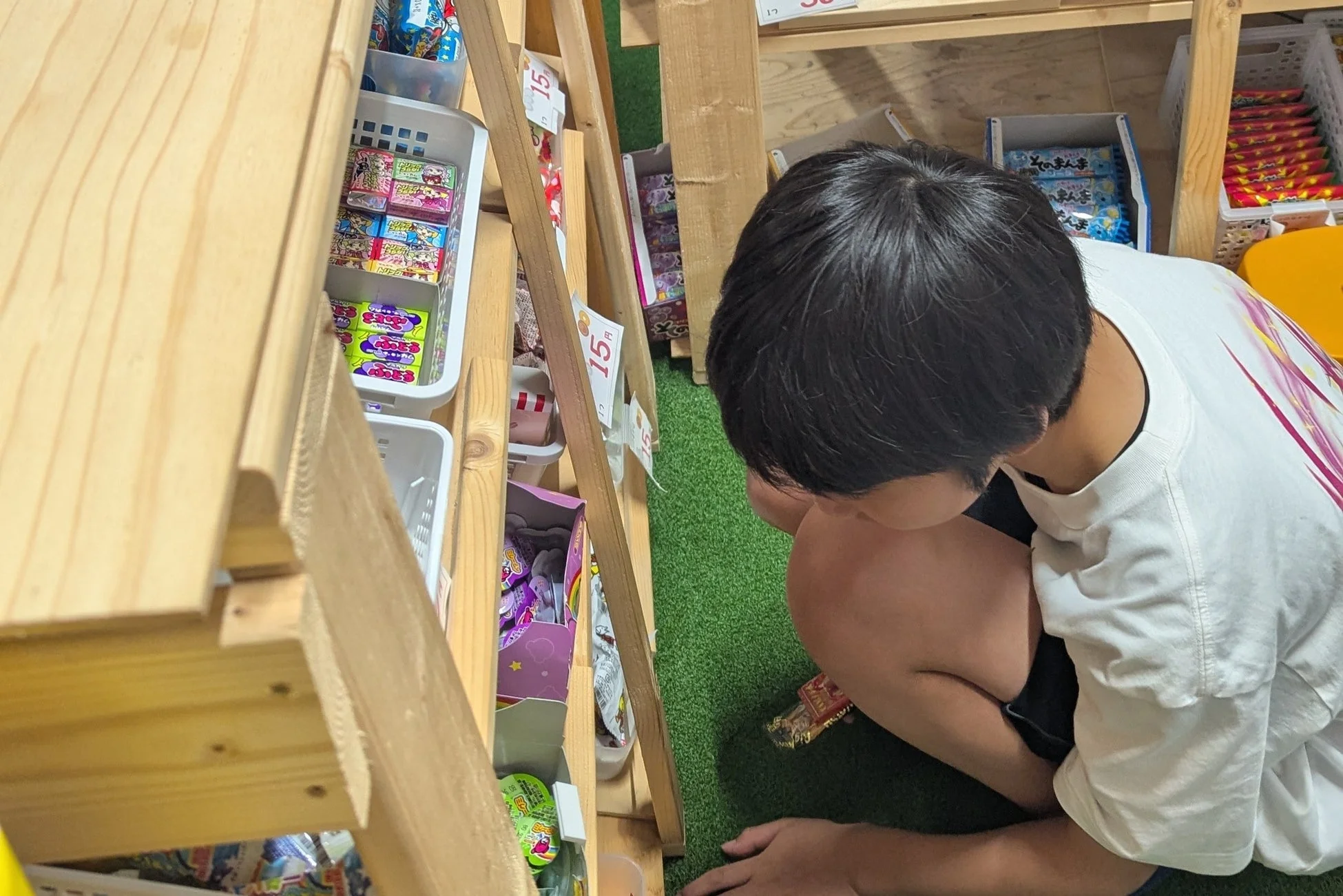 駄菓子屋のような店内で、少年がしゃがんで棚に並べられたカラフルなお菓子を熱心に選んでいる。15円の価格表示がいくつか見える。