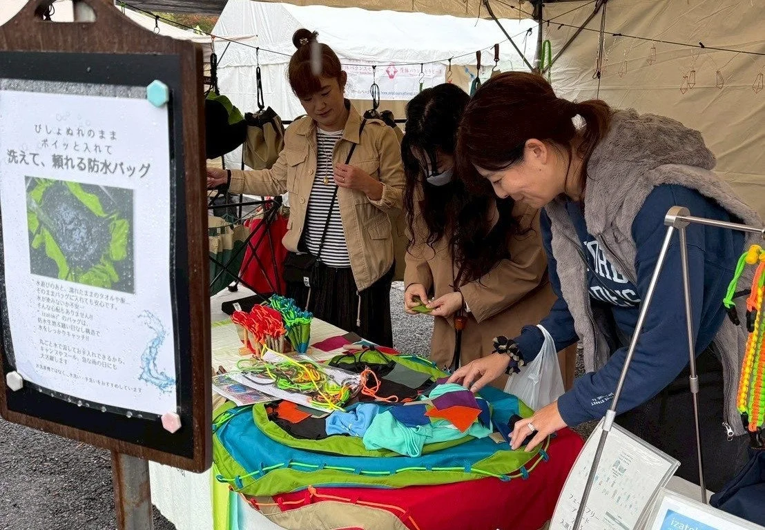 屋外のイベント会場で、3人の女性がカラフルな防水バッグやアウトドアグッズが並ぶ屋台で商品を見ています。