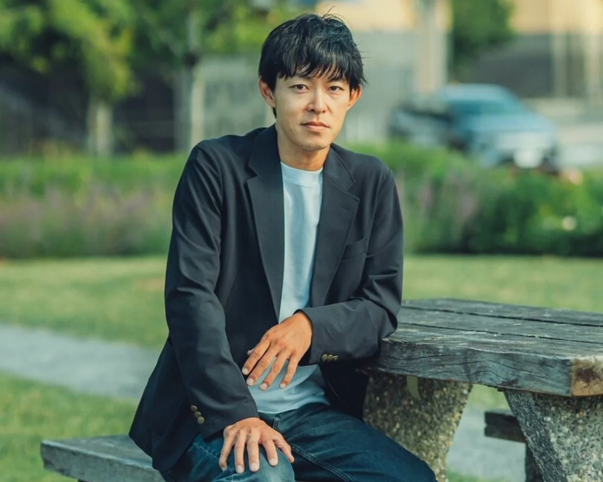 公園のような屋外で、黒いジャケットとジーンズを着用したアジア系の男性が木製のベンチに座っています。