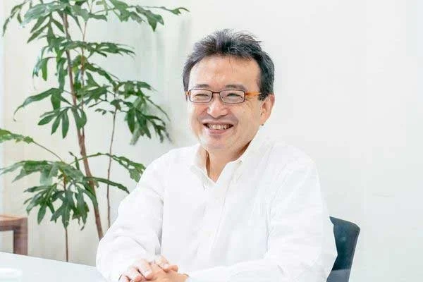 眼鏡をかけ白いシャツを着た中年の日本人男性が、室内でデスクに座り明るく微笑んでいます。背景には緑の植物が見えます。
