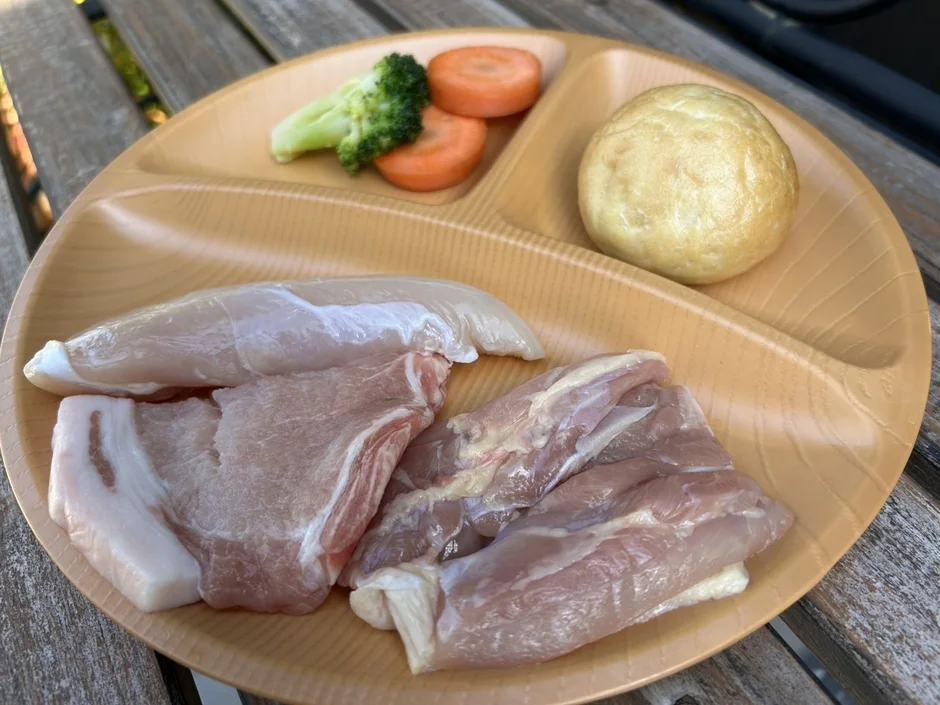木製のテーブルに置かれた仕切り付きプレートに、鶏むね肉、豚バラ肉、鶏もも肉などの生肉と、ブロッコリー、人参、パンが盛り付けられています。屋外での食事準備の様子です。