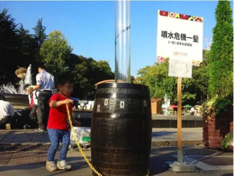 青空の下、公園のような場所で、一人の子供が大きな樽の横に立っています