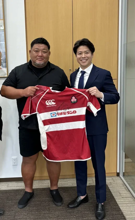 二人の男性が笑顔で、サイン入りのラグビー日本代表ジャージを掲げている記念写真です。