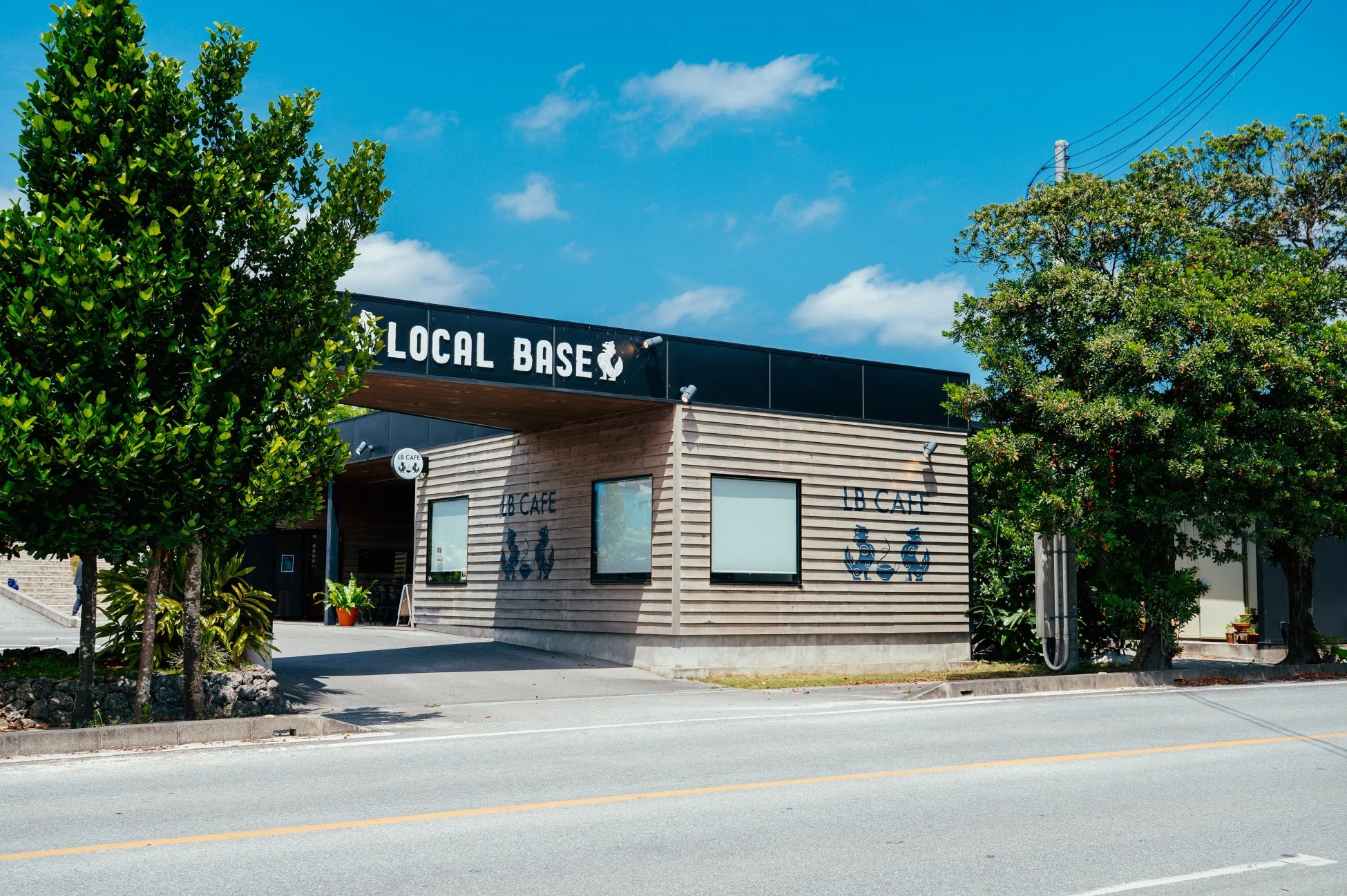 青い空の下、木製の外壁と黒い看板が特徴的な「LOCAL BASE」という名の建物。側面には「LB CAFE」の文字と猫のロゴがあり、カフェのようです。