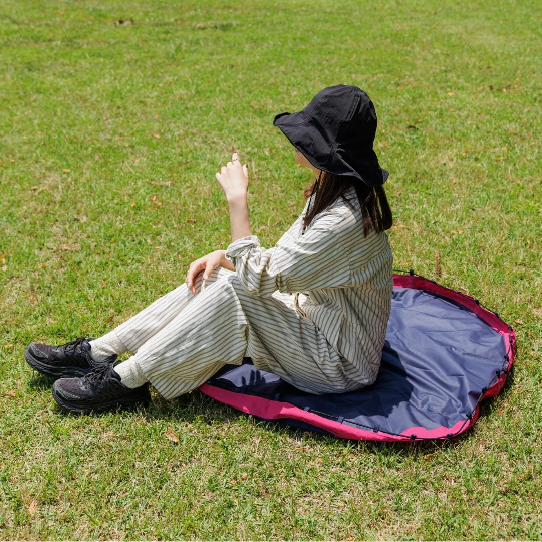 緑の芝生の上で、黒い帽子とストライプの服を着た女性がレジャーシートに座り、遠くを指差しています。