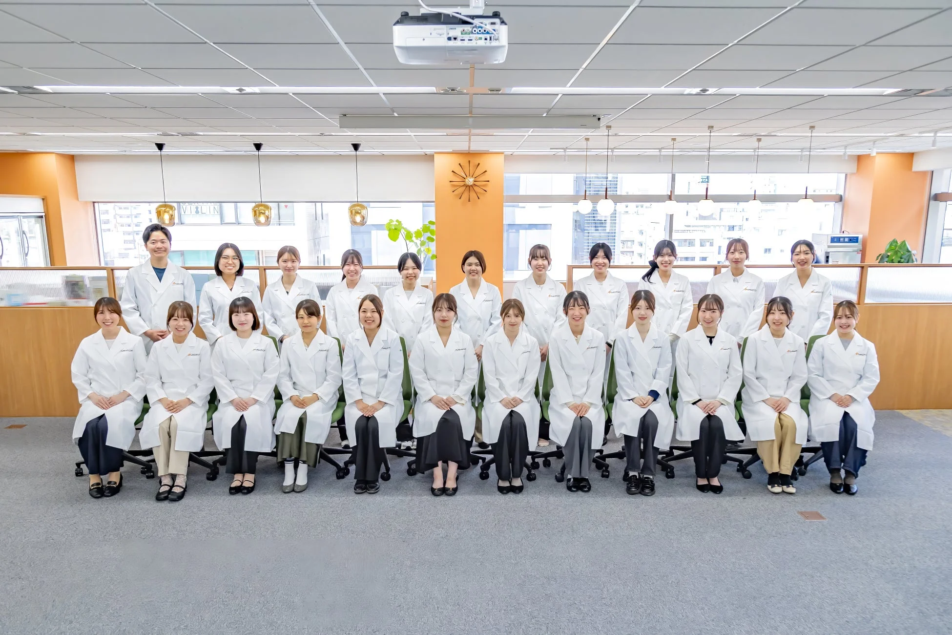 明るいオフィス空間で、白衣を着用した20人以上の男女が集合写真を撮影している様子