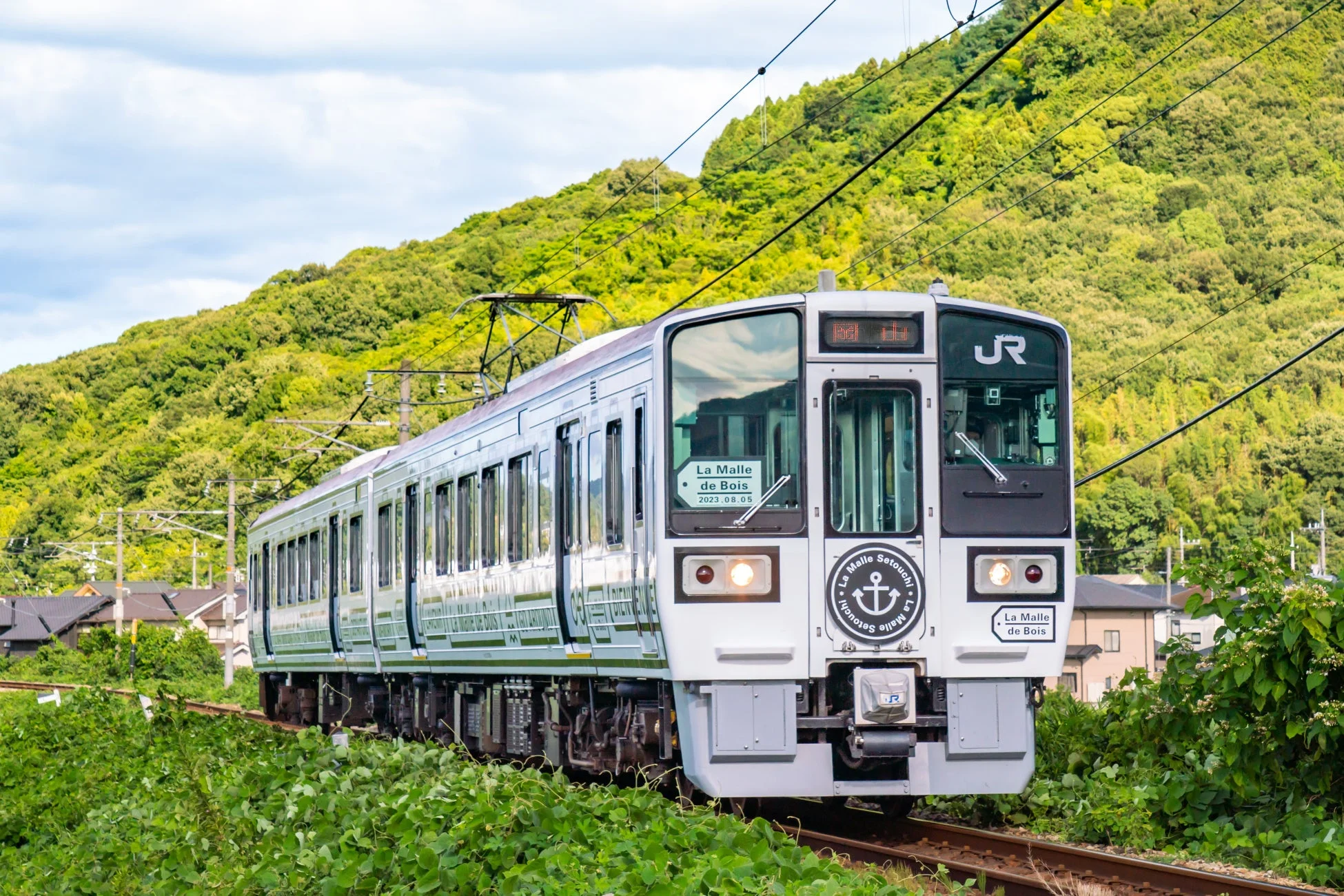 緑豊かな山々を背景に、白い車体に錨のマークが特徴的な観光列車「La Malle de Bois」が線路を走行している風景。