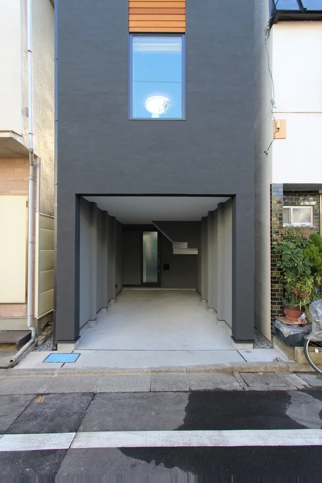 都市部に建つ、ダークグレーの外壁と木目調のアクセントが特徴的なモダンな狭小住宅です。1階部分はビルトインガレージとなっており、奥に玄関が見えます。