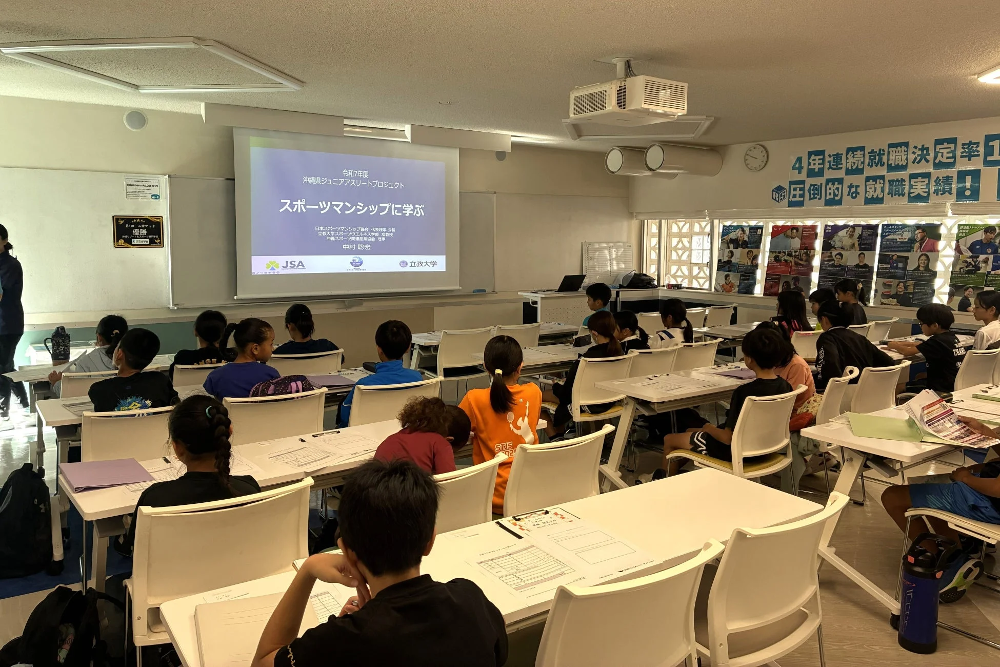 教室で多数の子供たちが「沖縄県ジュニアアスリートプロジェクト」の一環として「スポーツマンシップに学ぶ」というテーマの授業を受けている様子