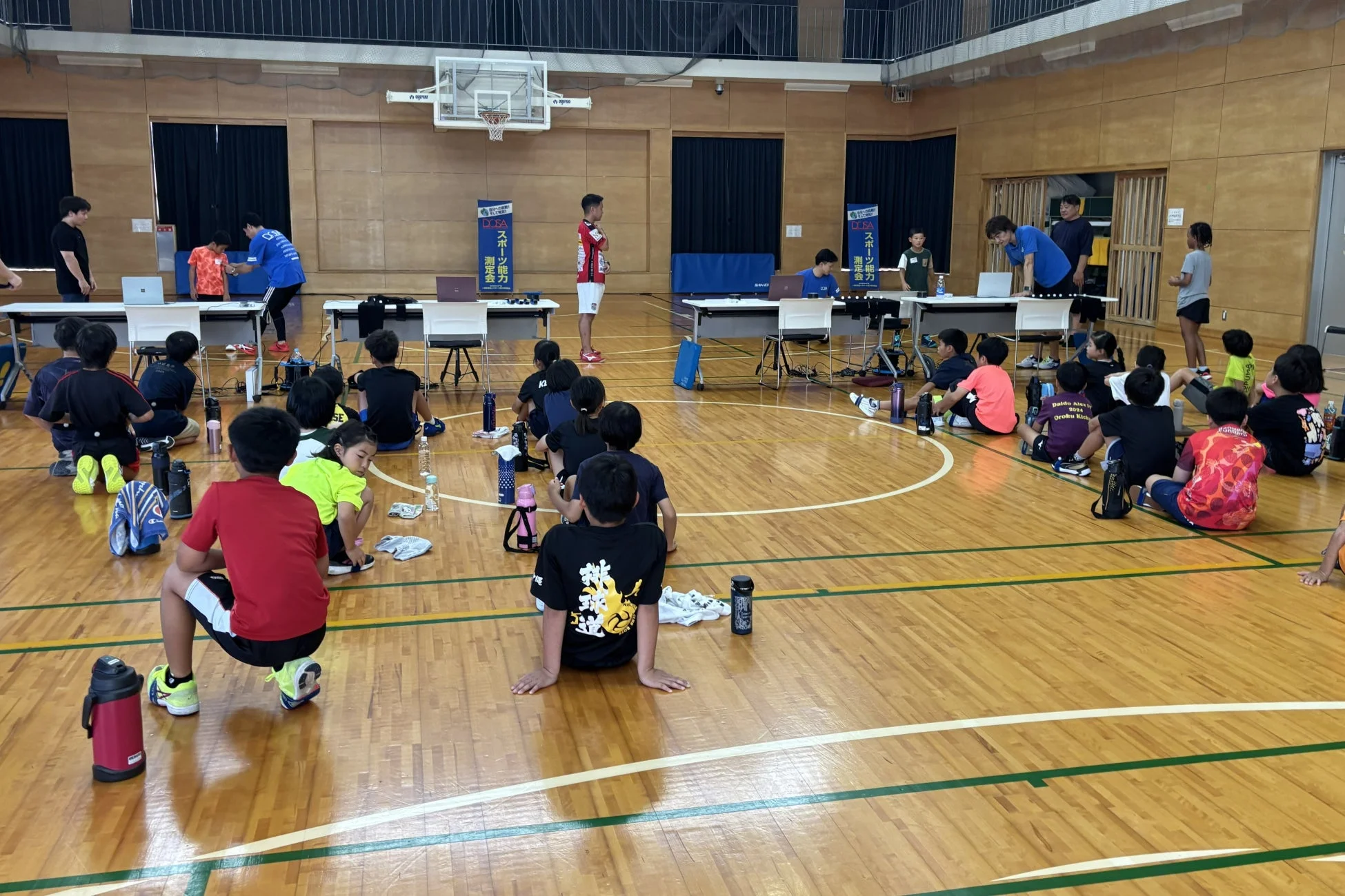 体育館でスポーツ能力測定会が開催され、多くの子どもたちが説明を聞いている様子