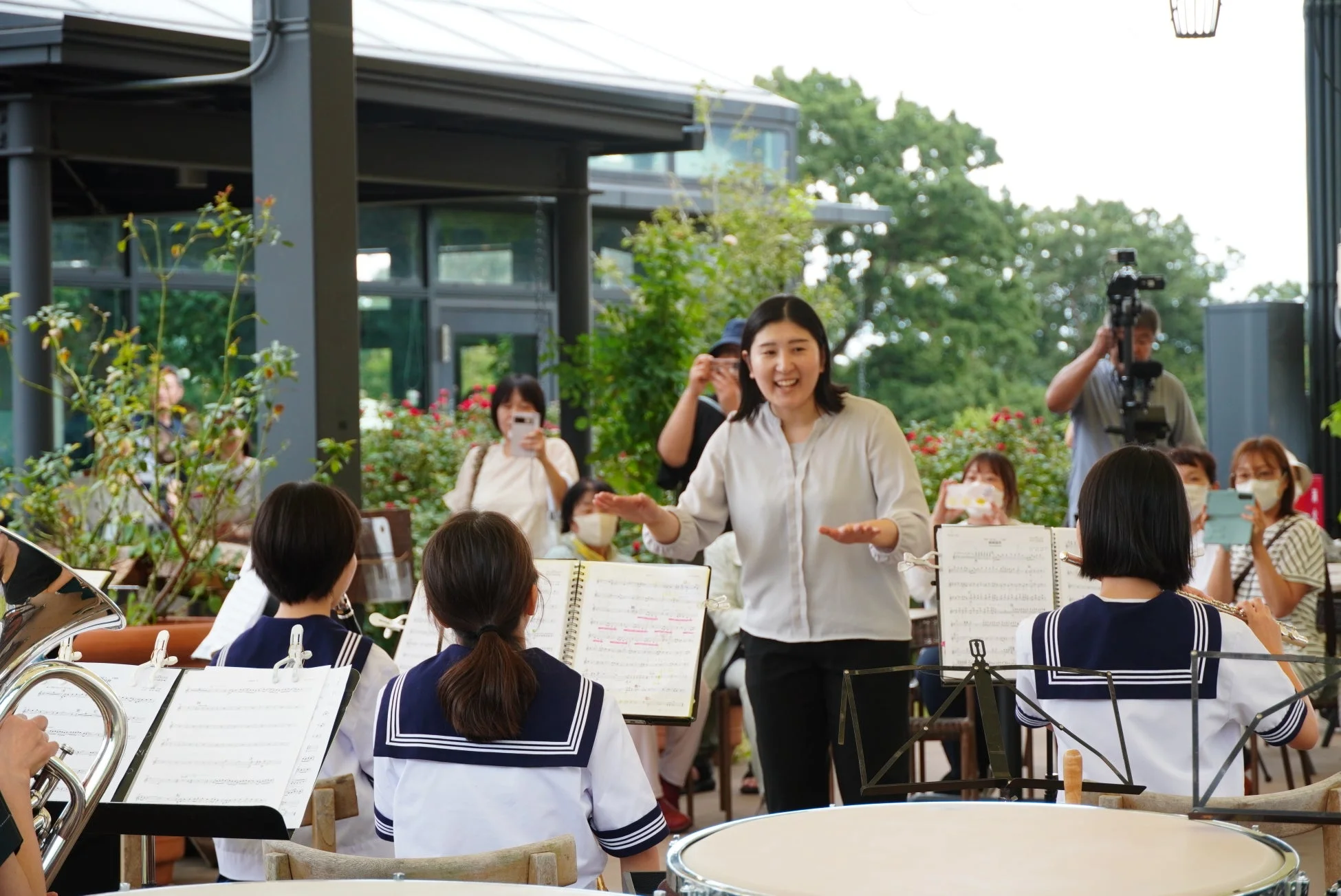 屋外の会場で、制服を着た学生たちが吹奏楽の演奏をしており、一人の女性が笑顔で指揮をしています