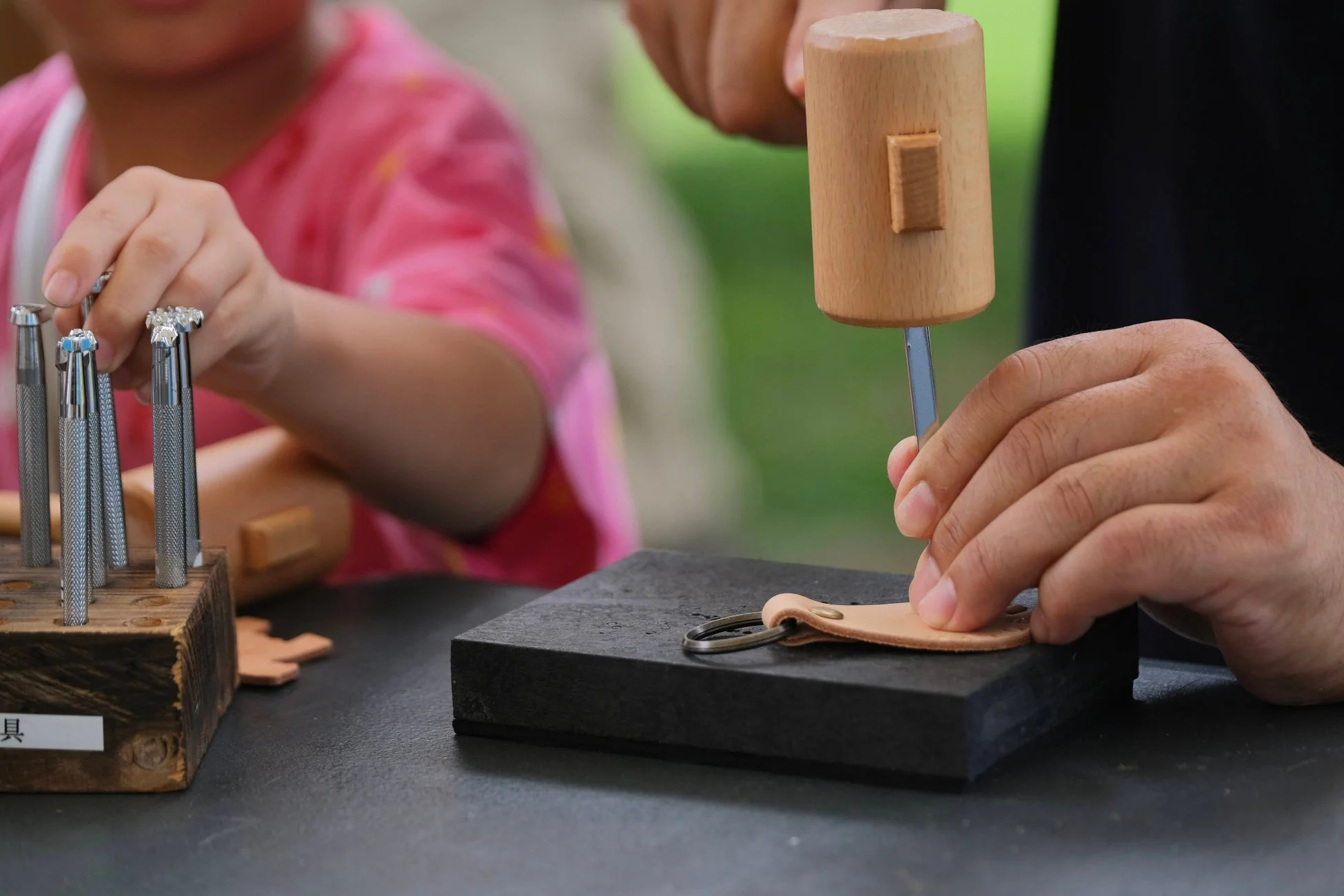 大人が木槌で革製品に刻印を施し、子供がレザークラフト用の工具を選んでいる様子