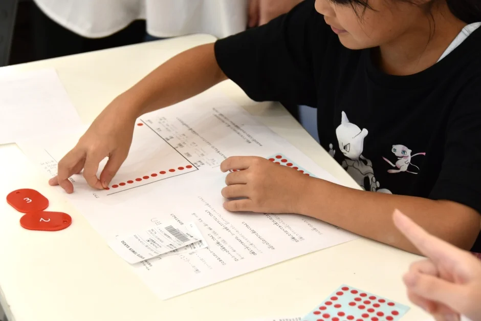 机に向かい、赤いドットが描かれたワークシートと数字の教材を使って学習する子供の様子