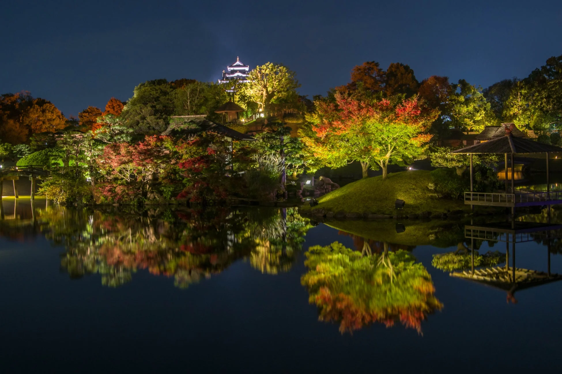 ライトアップされた日本庭園の夜景で、色鮮やかな紅葉の木々や奥に見える城が池に美しく映り込んでいます。