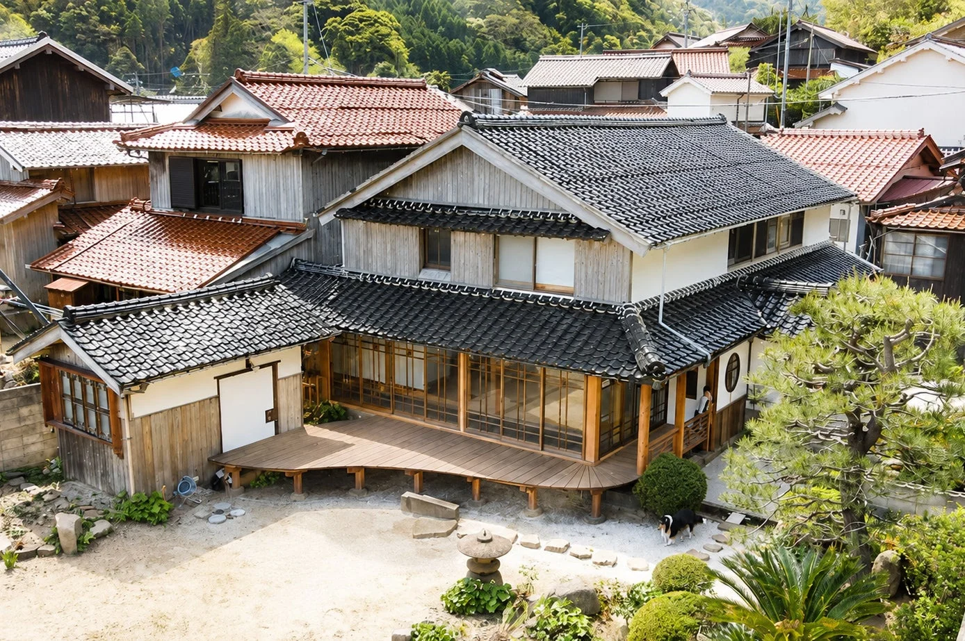 緑豊かな山を背景に、黒と赤の瓦屋根が特徴的な日本の伝統的な家屋が立ち並ぶ集落の風景です。手前には庭園があり、石灯籠や犬の姿も見え、穏やかな雰囲気を醸し出しています。