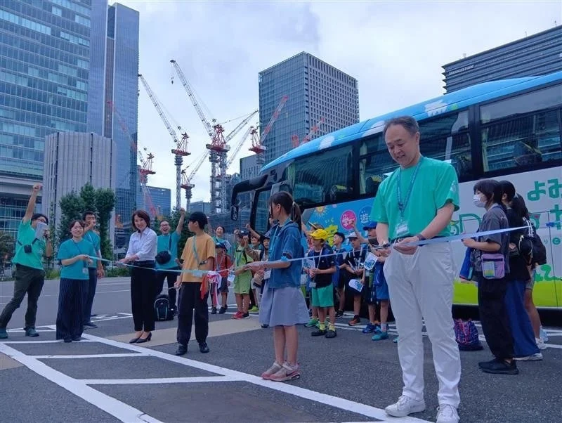 都市の高層ビルと建設クレーンを背景に、大人と子供たちがテープカットセレモニーに参加しています。青いバスが隣接し、新しいイベントの開始を祝う賑やかな様子が描かれています。
