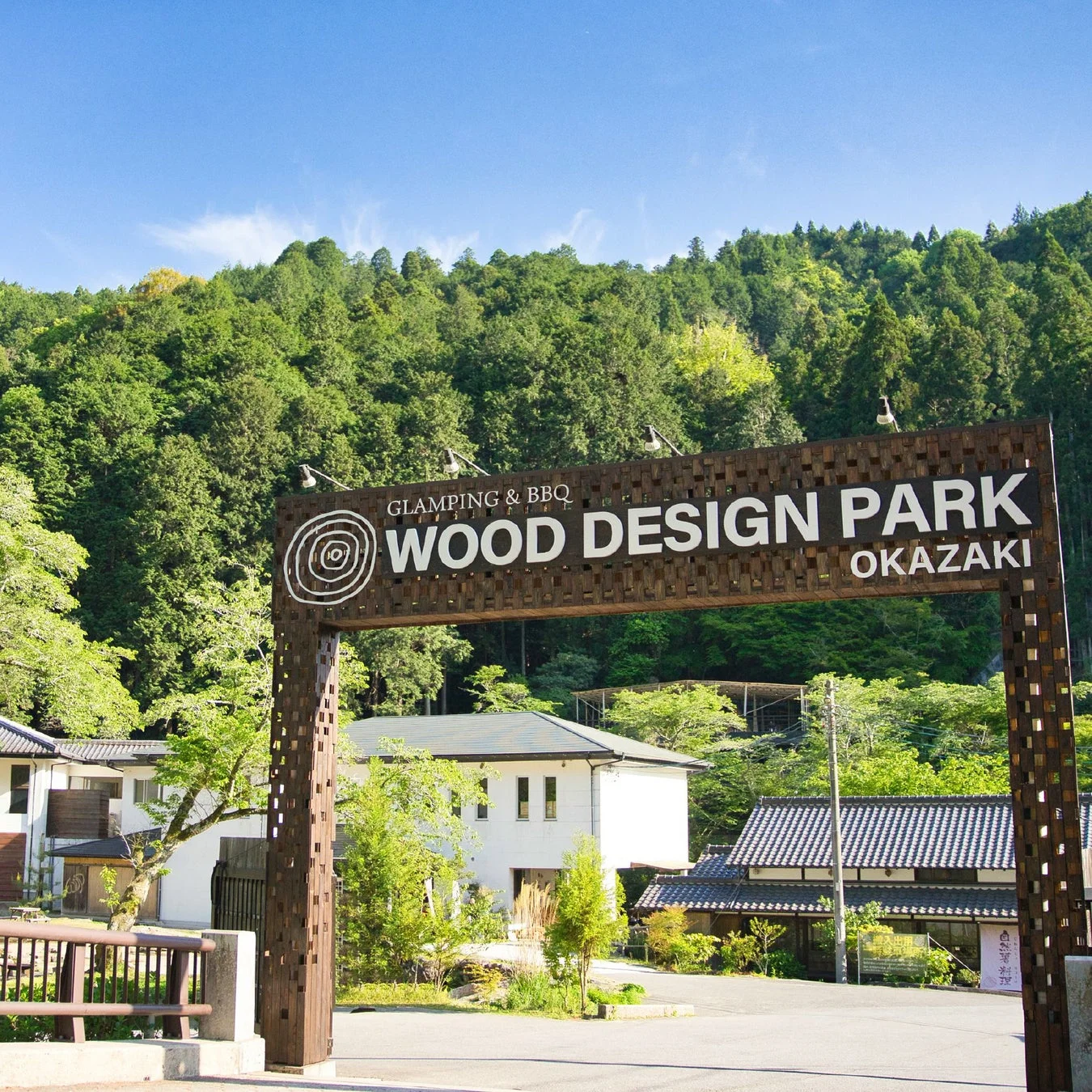GLAMPING & BBQ WOOD DESIGN PARK OKAZAKIと書かれた木製の看板が目立つ、自然豊かな施設のエントランス風景