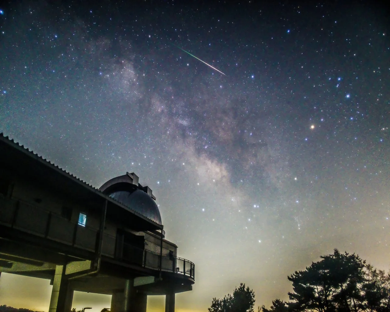 満天の星が輝く夜空に天の川と流星が見え、手前にはドーム型の観測所と木々が写っています。