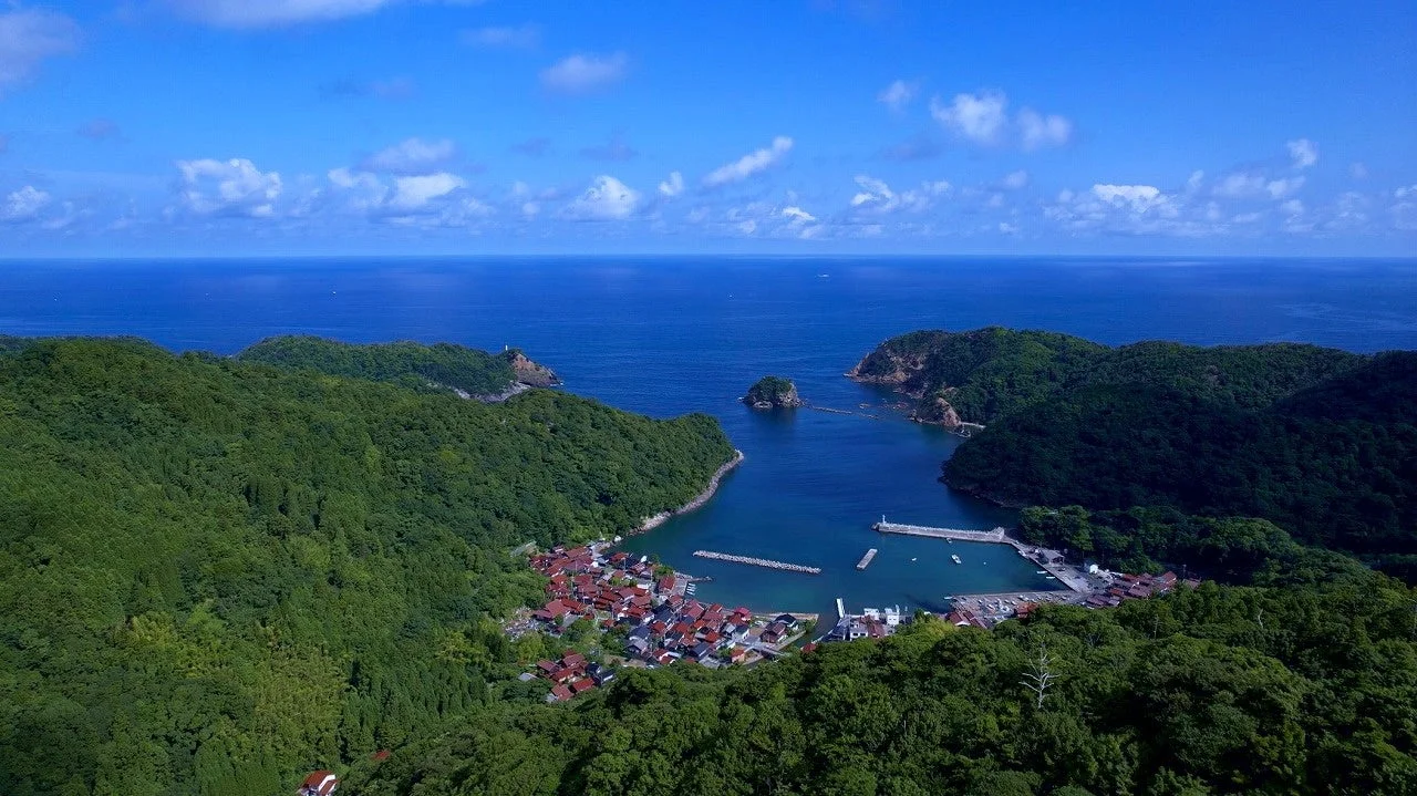 緑豊かな山々に囲まれた湾に広がる小さな集落を空から捉えた美しい風景写真です。紺碧の海と青い空、白い雲が広がり、穏やかな港には防波堤が見えます。