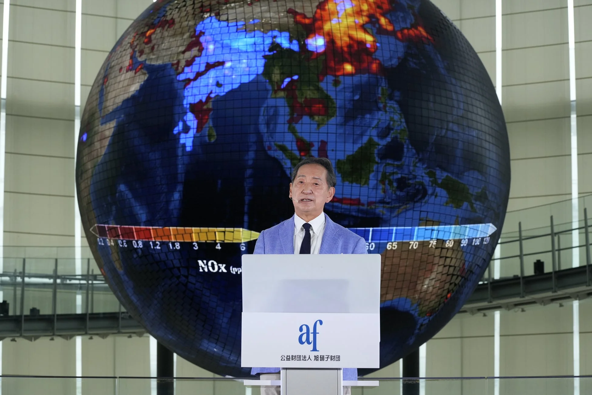 男性が壇上で講演しており、背後にはNOx濃度などの環境データを示す巨大な地球儀のディスプレイが映し出されています。演台には「公益財団法人 旭硝子財団」のロゴが見え、環境に関する発表が行われている様子です。