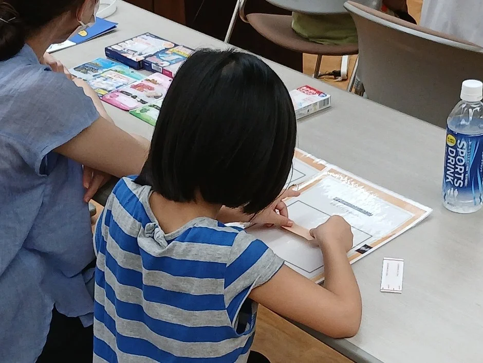 机に向かい、紙を使った作業に集中している子供と、その隣に座る大人の様子