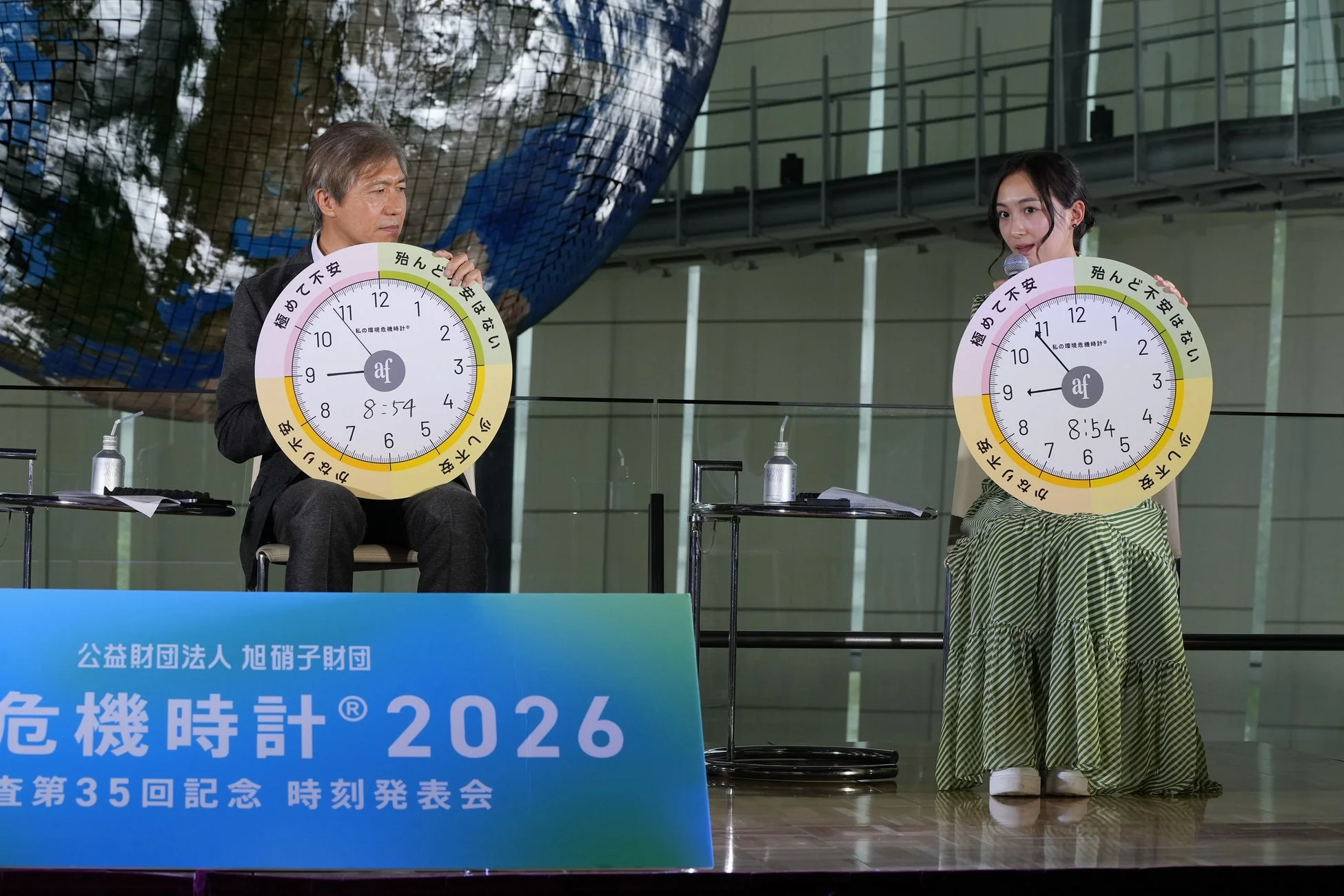 旭硝子財団による「危機時計® 2026」第35回記念時刻発表会の様子。男女2名が環境危機時計を持ち、時刻は8時54分を示している。時計の文字盤には環境への不安度が示され、背後には地球儀が見える。