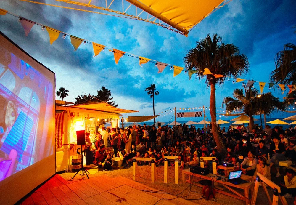 夕暮れ時、ビーチのような場所で屋外スクリーンに映し出された映画を鑑賞する人々