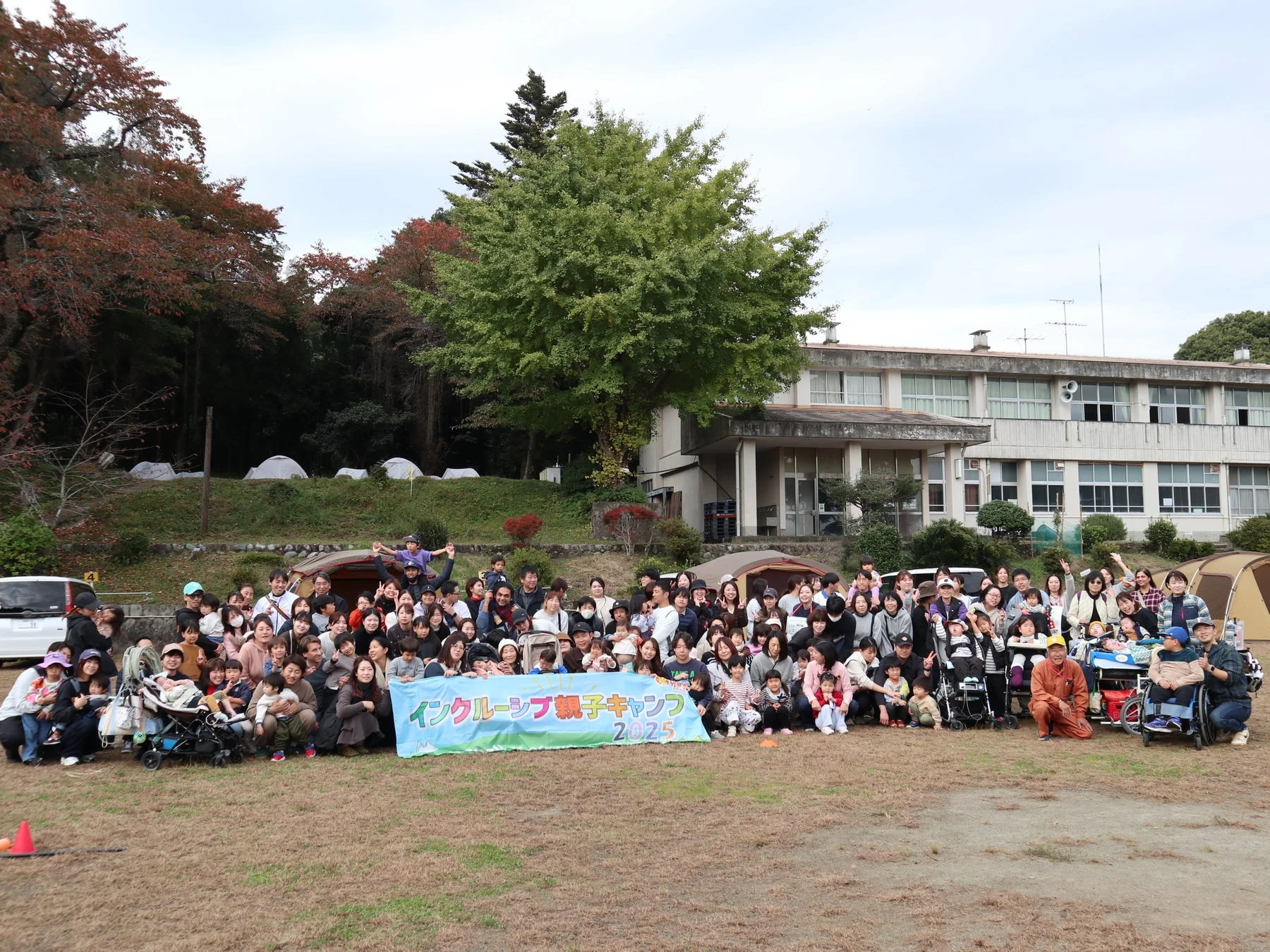 「インクルーシブ親子キャンプ 2025」と書かれた横断幕の前で、大人と子供が大勢集まり笑顔で写る集合写真